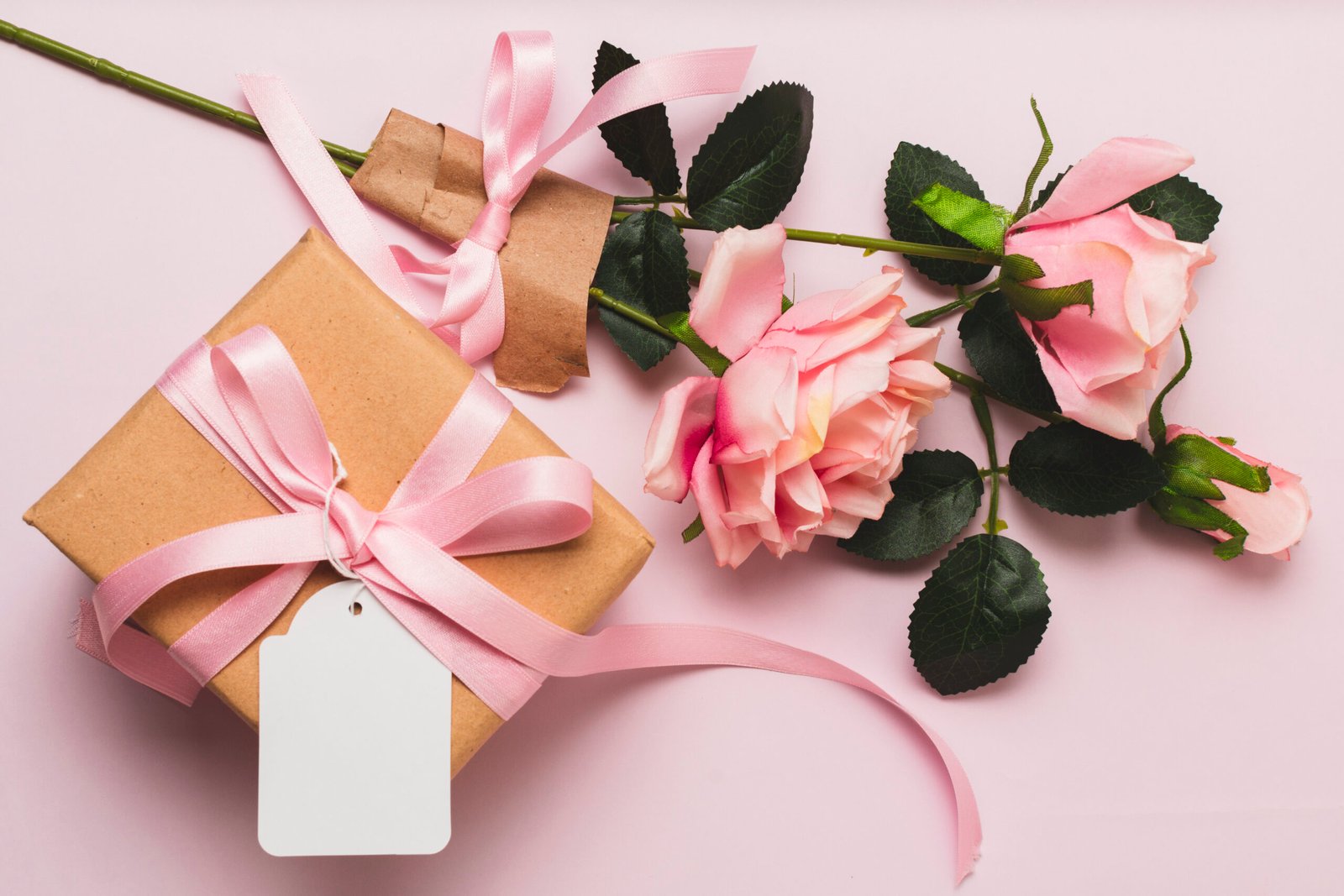 Gift box wrapped in brown paper with a pink ribbon, placed next to a bouquet of pink roses on a soft pink background.